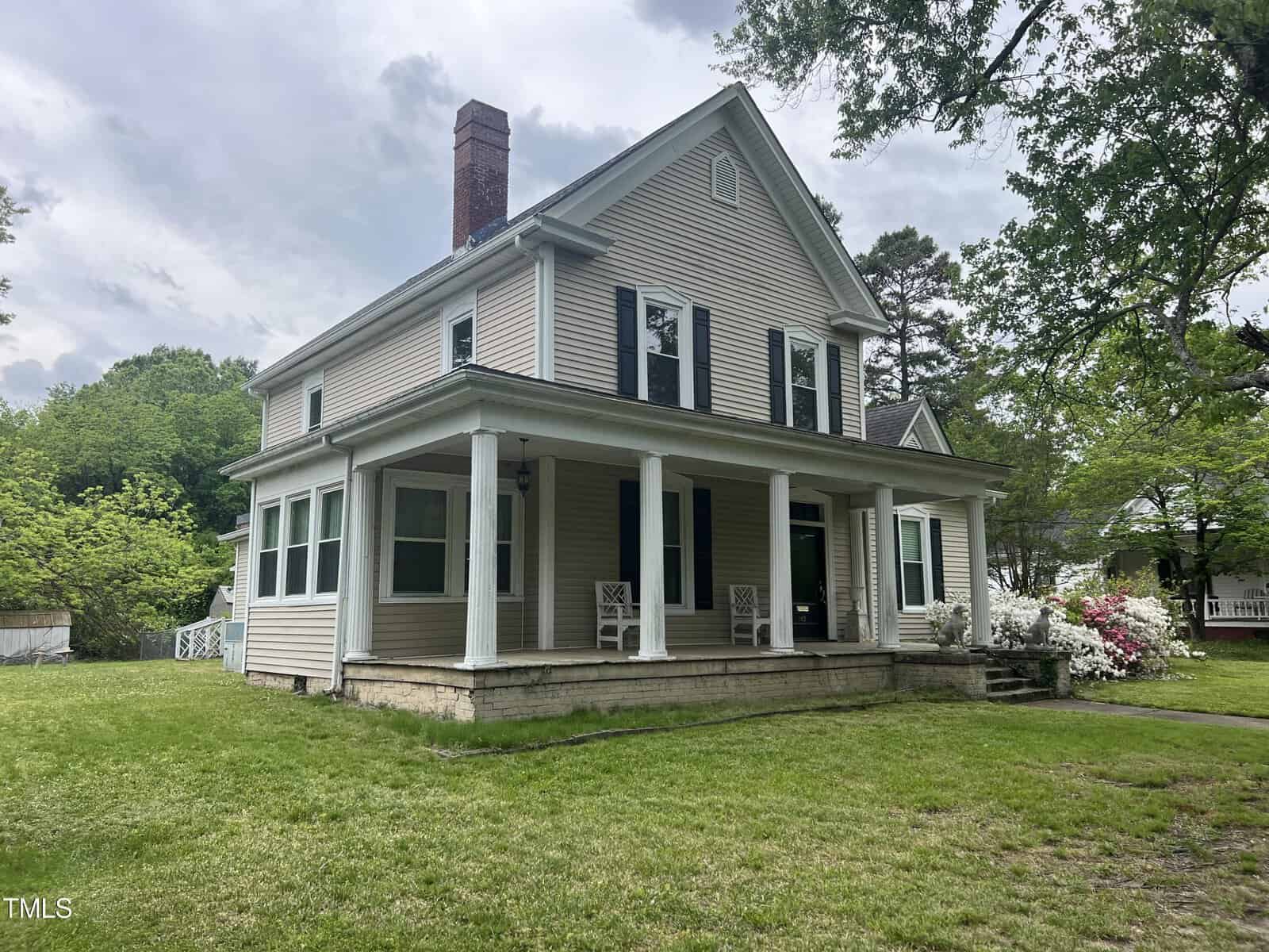 A two-story, beige colonial house with white columns and a chimney, surrounded by a green lawn and trees. A porch with chairs is visible, and the sky above is cloudy.
