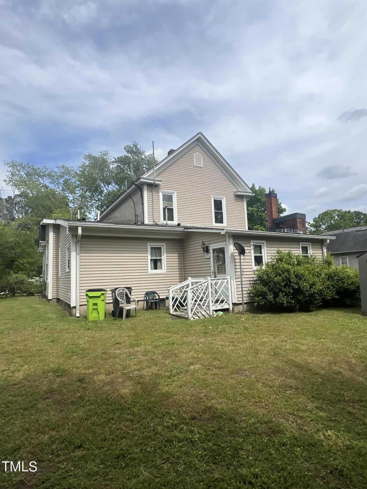A beige two-story house with a small white patio and steps, surrounded by a grassy yard. There are two green trash bins and a few outdoor chairs near the patio. Trees and shrubs are in the background under a cloudy sky.