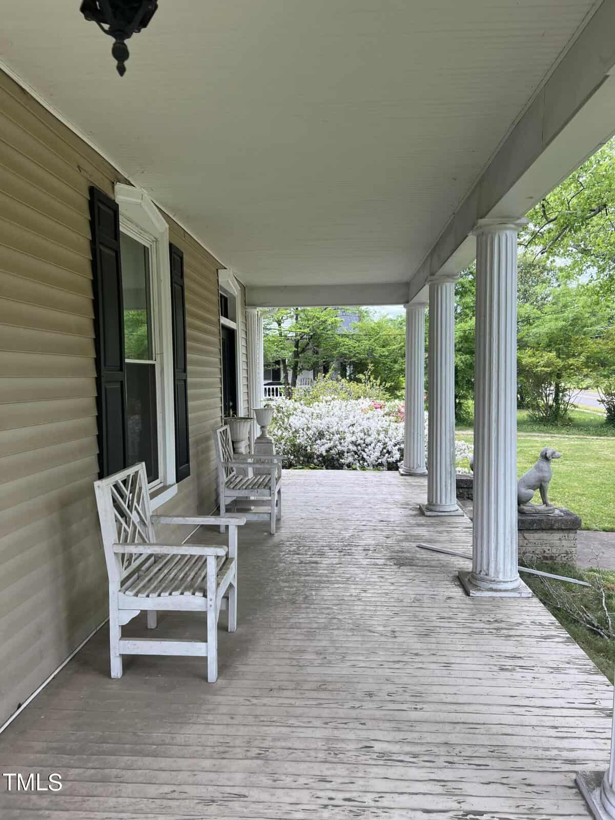 A long, covered porch with two white wooden chairs and a small table. The area is surrounded by greenery, including bushes with white flowers. A decorative stone dog is perched at the edge of the porch.