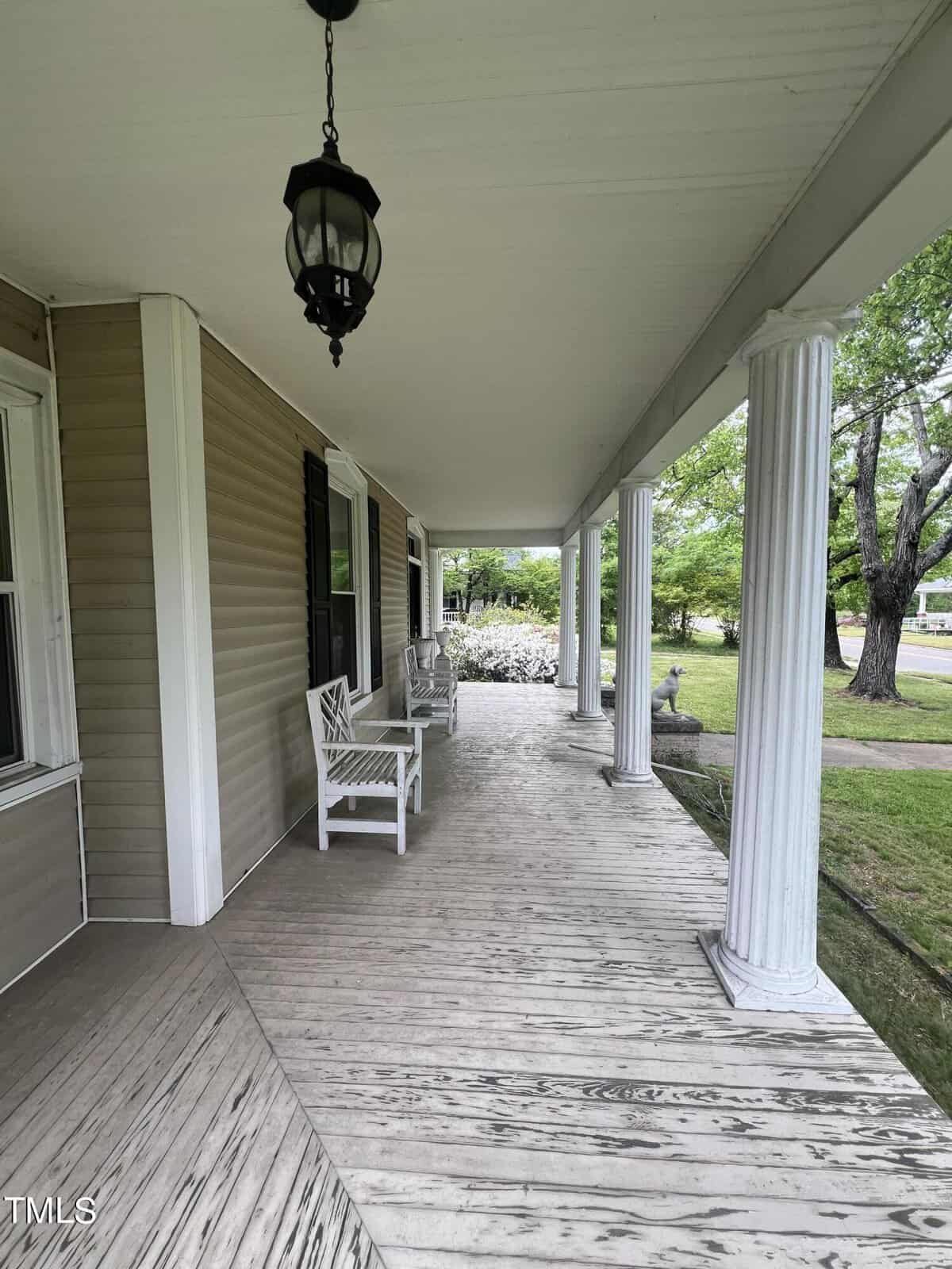 A spacious wooden porch with white columns and two white chairs. A black hanging lantern is on the ceiling. The porch overlooks a lush green lawn with bushes and a tree in the background.