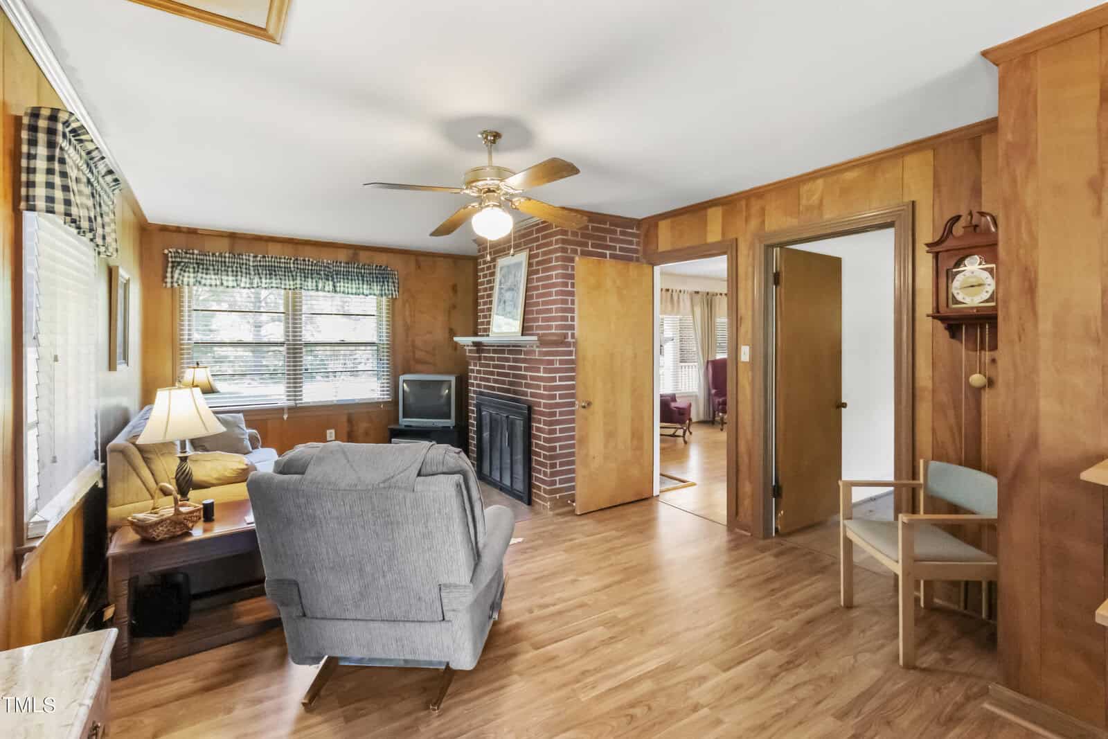 A cozy living room featuring a brick fireplace, wooden paneling, hardwood floors, and furnishings such as a grey armchair, lamp, and wall-mounted clock. natural light streams in through a window.