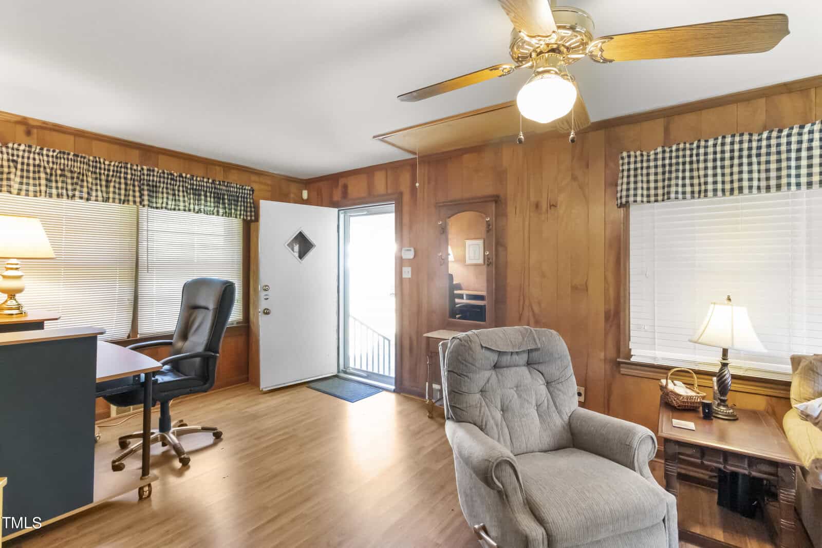 A cozy home office with wooden panel walls, featuring a desk with a black chair, a recliner, and a lamp. there's a ceiling fan above and a door leading outside with natural light streaming in.