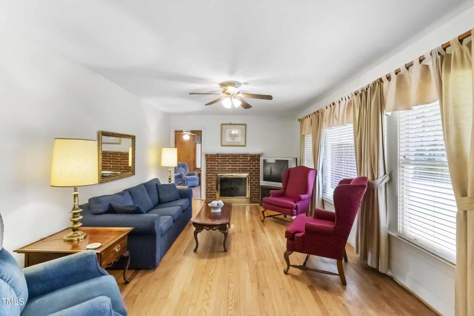 A cozy living room featuring two blue sofas, two red armchairs, a wooden coffee table, a fireplace, and soft lighting from table lamps. hardwood floors and light curtains complete the homely space.