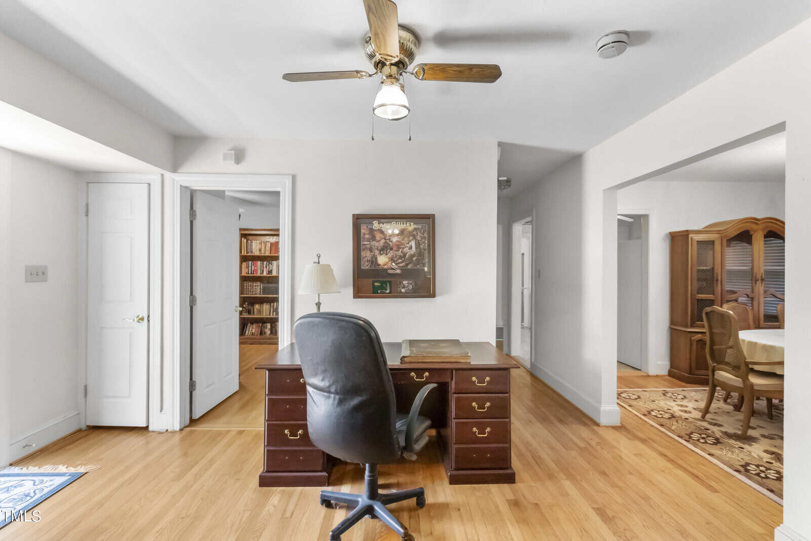 A spacious home office featuring a large wooden desk with a black office chair, a ceiling fan above, and an open doorway leading to a dining room. bright walls and carpeted floor enhance the room's airy feel.