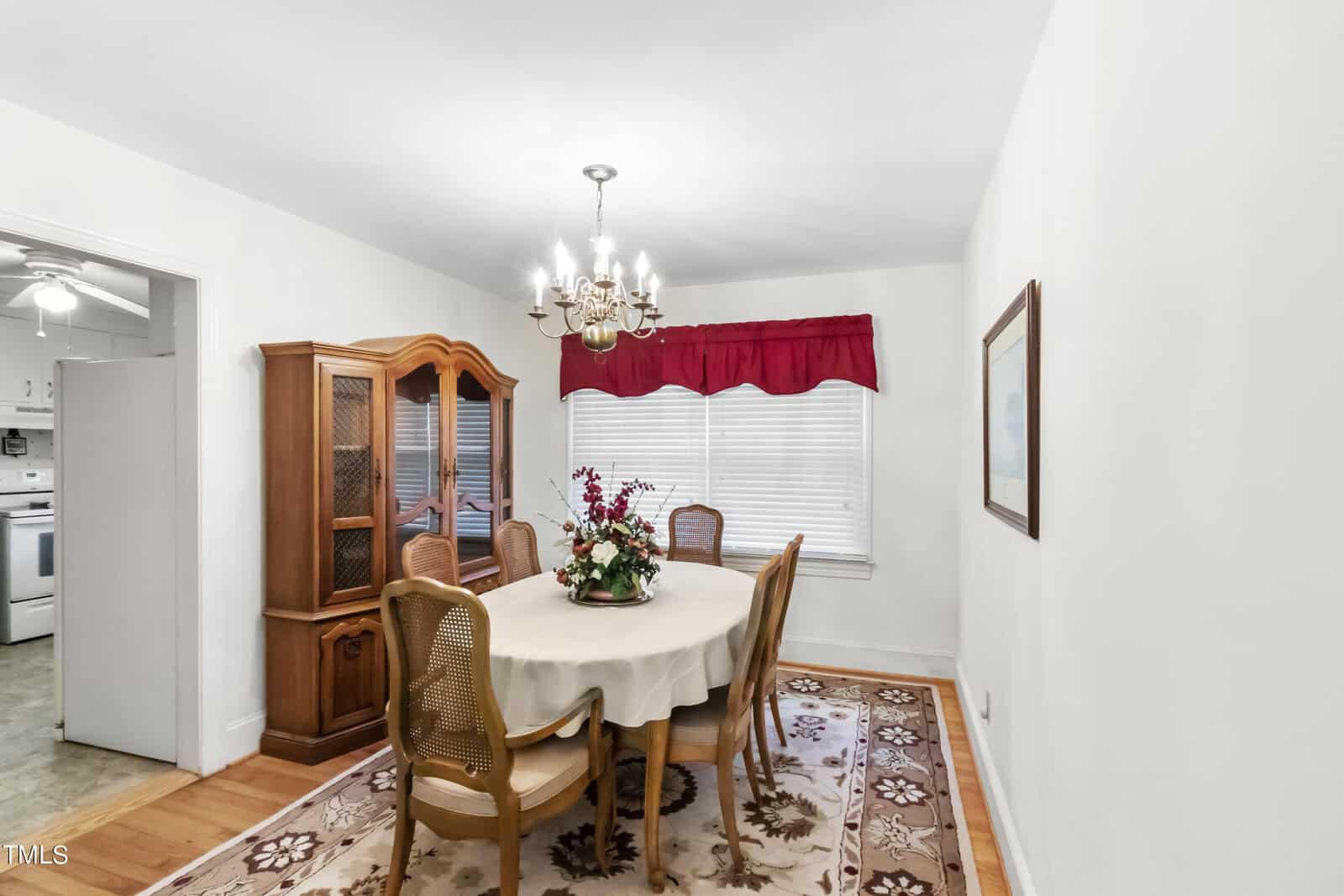 A bright, well-lit dining room with a wooden dining table set for four, an elegant chandelier above, and a hutch to the left. a kitchen is visible in the background. decor includes a red valance and a floral centerpiece.