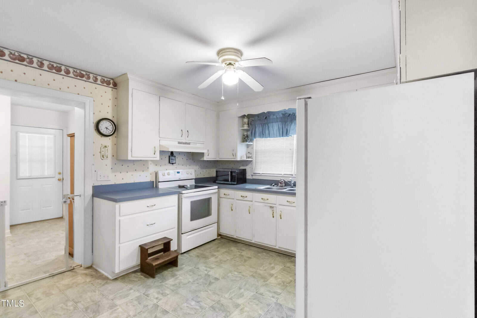 A small, tidy kitchen with white cabinets, blue countertops, and white appliances. a ceiling fan is above, and there is a window with a blue curtain to the right.