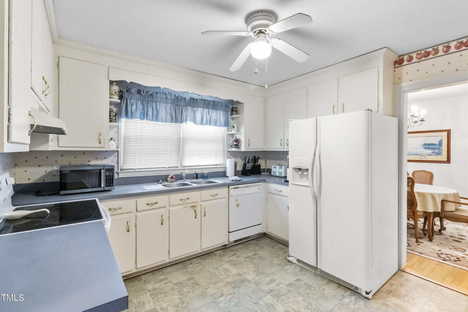 A vintage-style kitchen with white cabinets, a large refrigerator, a ceiling fan, and a window with a blue curtain. there's a microwave on the counter and a dining area visible in the background.