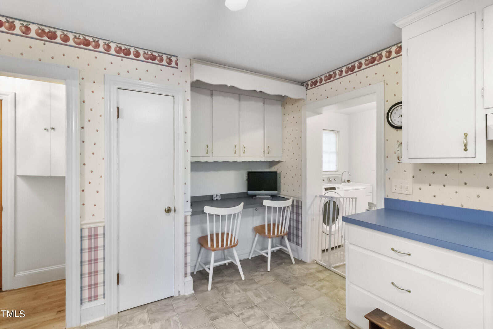 A quaint kitchen with blue countertops, white cabinets, and a small dining area with a round table and two chairs. the room features polka-dot wallpaper with a fruit-themed border.