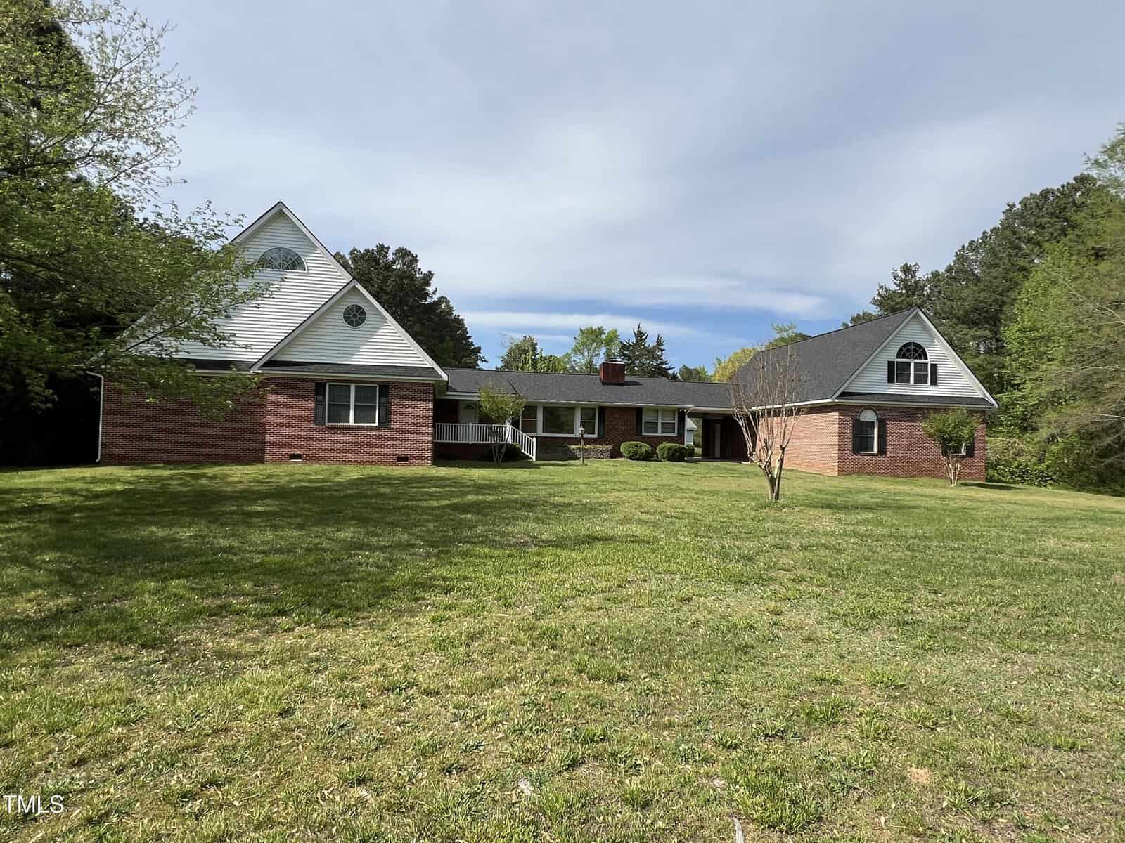 A spacious brick house with a prominent white gabled roof, nestled in a large grassy yard surrounded by trees under a clear blue sky.