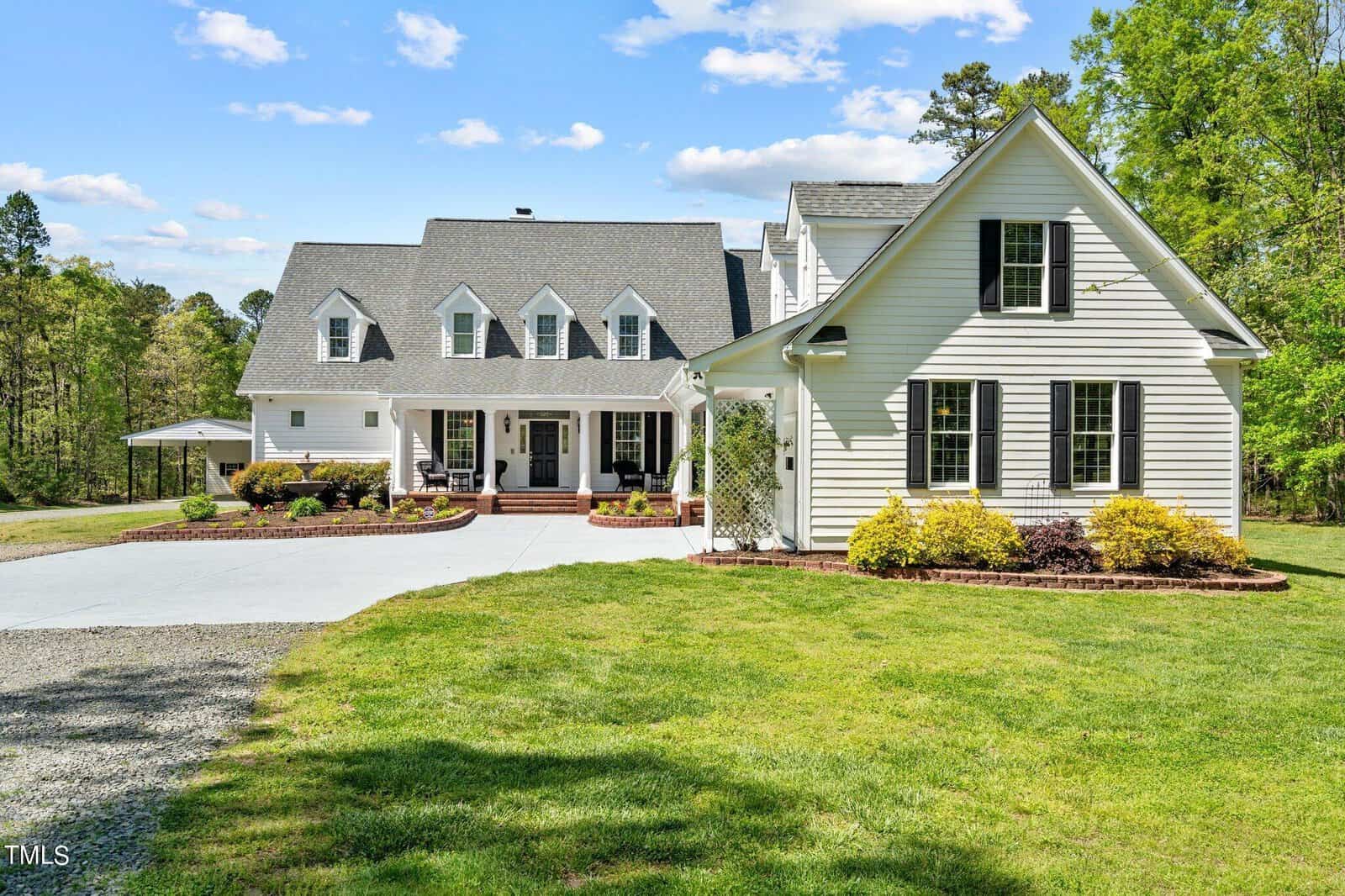 A large white two-story house on 3195 Range Road boasts a gray roof, multiple gables, and a covered front porch. The gravel driveway gracefully leads to a landscaped yard with shrubs and trees in the background. It's a bright, sunny day with just a few clouds adorning the sky.