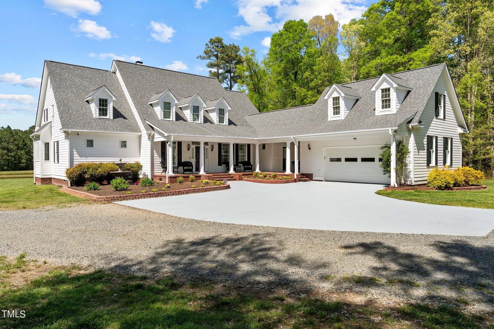 A large, two-story white house with a gray roof and multiple dormer windows, surrounded by greenery. It features a wrap-around porch and a double garage. The sky is clear with a few clouds. A gravel driveway leads to the house.
