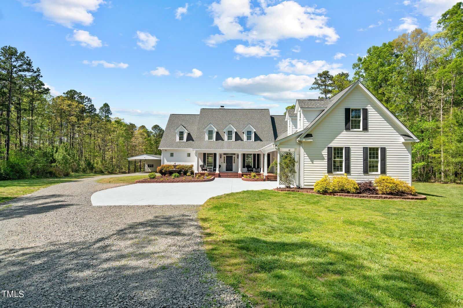 A large, two-story white house with a gray roof, surrounded by green grass and trees. There is a gravel driveway leading to the entrance and a wooden porch at the front. The sky is blue with scattered clouds.