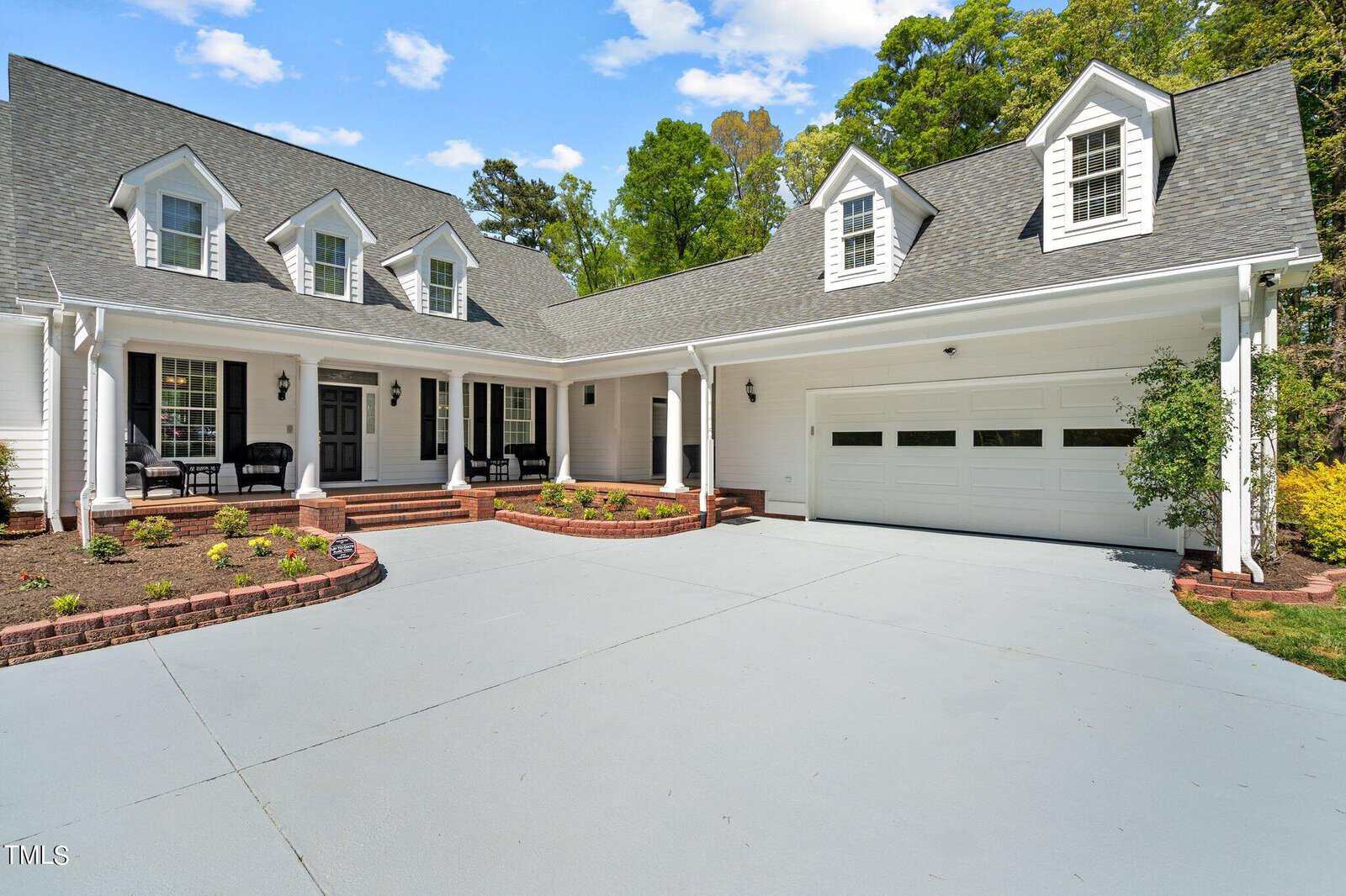 A spacious white house features a sloped roof with dormer windows, a welcoming front porch, and a two-car garage. The driveway is expansive, and there are well-maintained shrubs and trees surrounding the property.