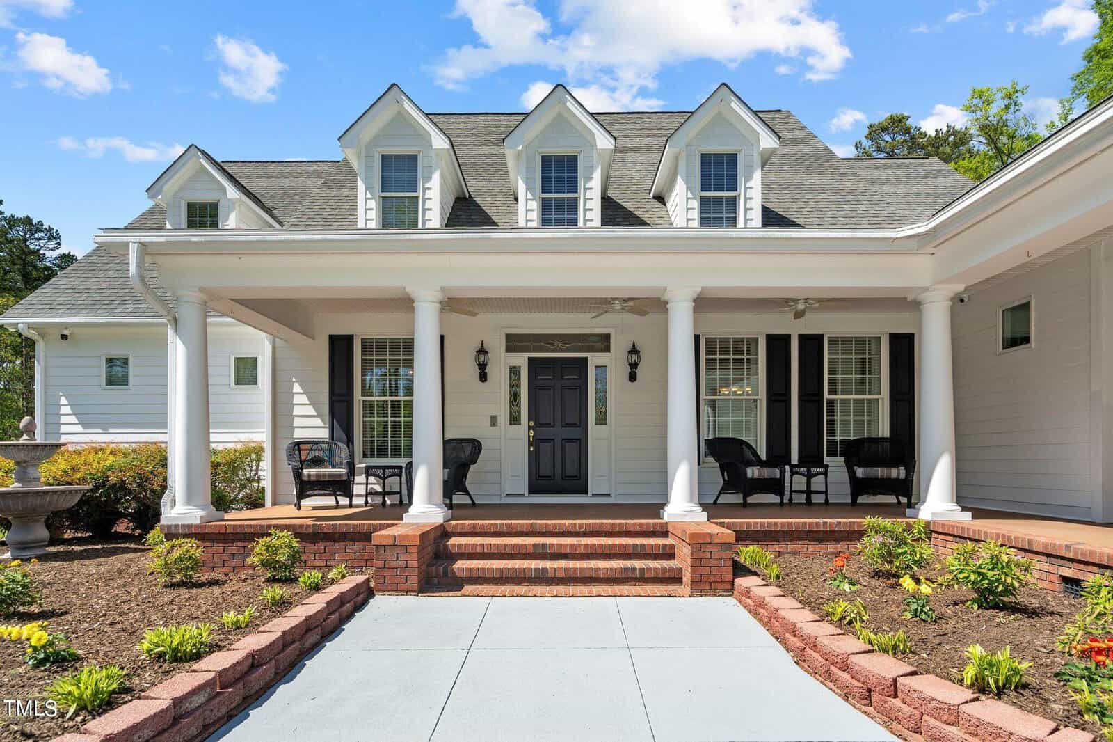 A white two-story house with a gray roof, featuring a covered front porch with brick steps. The porch has chairs and decorative columns. A landscaped garden with small plants and a decorative urn is in the foreground under a partly cloudy sky.