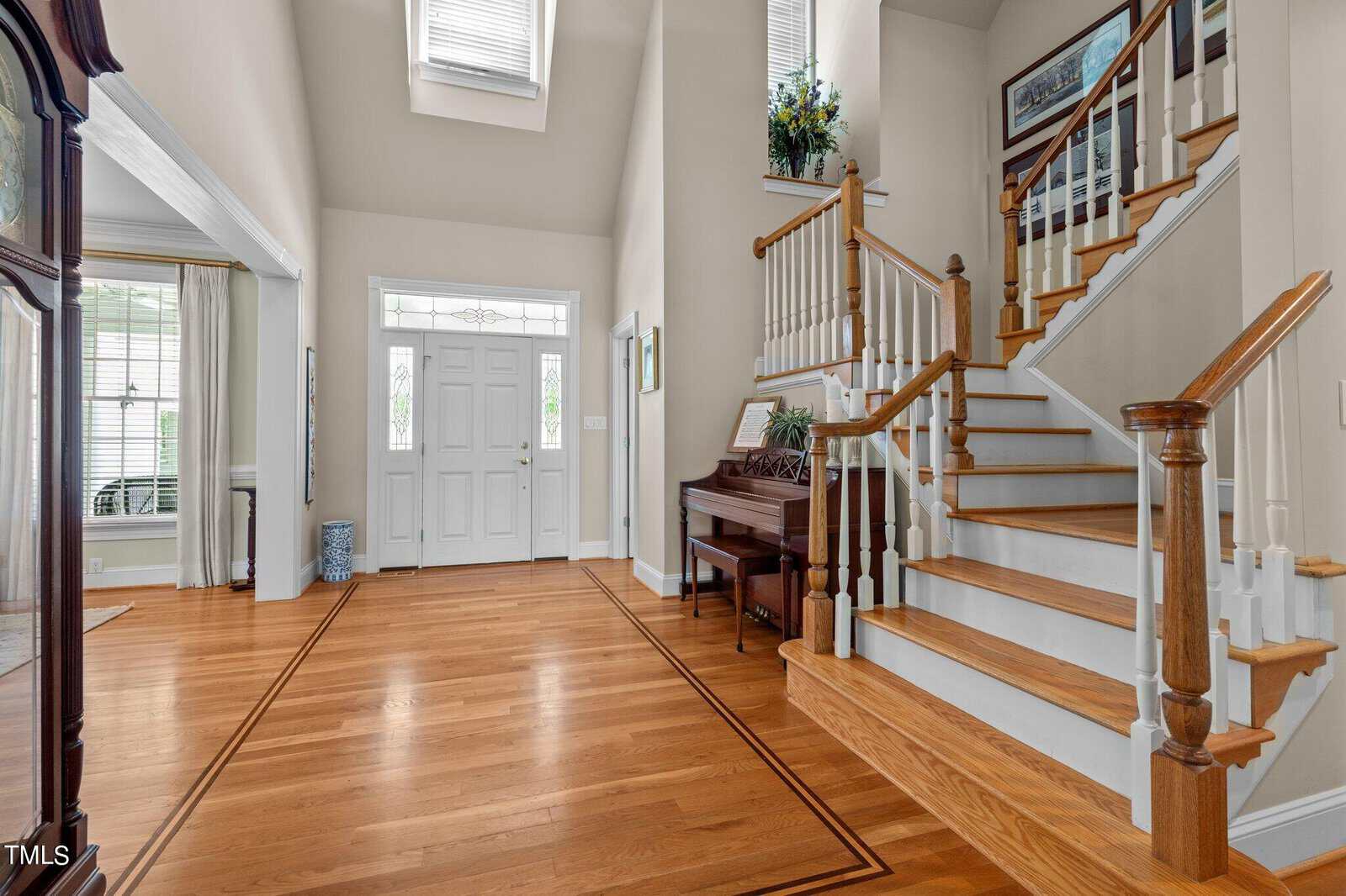 A spacious, well-lit foyer with wooden floors, white walls, and a grand staircase. A piano is positioned near the staircase, with a plant on top. Large windows and a decorative glass door let in natural light.