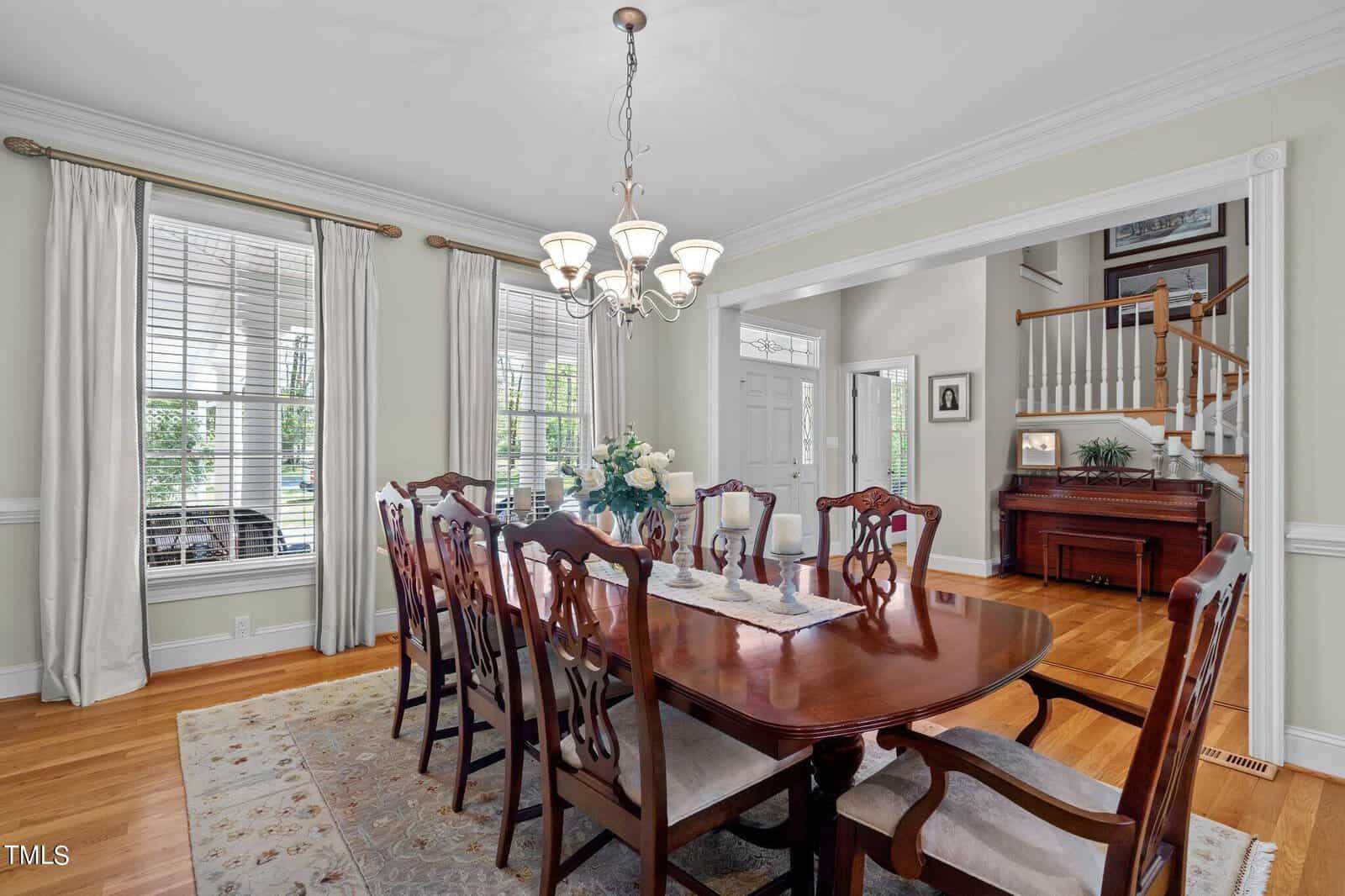 A formal dining room with a dark wood table and eight matching chairs. A chandelier hangs above the table, which has a floral centerpiece. Large windows with curtains line the wall. A staircase and a piano are visible in the background.