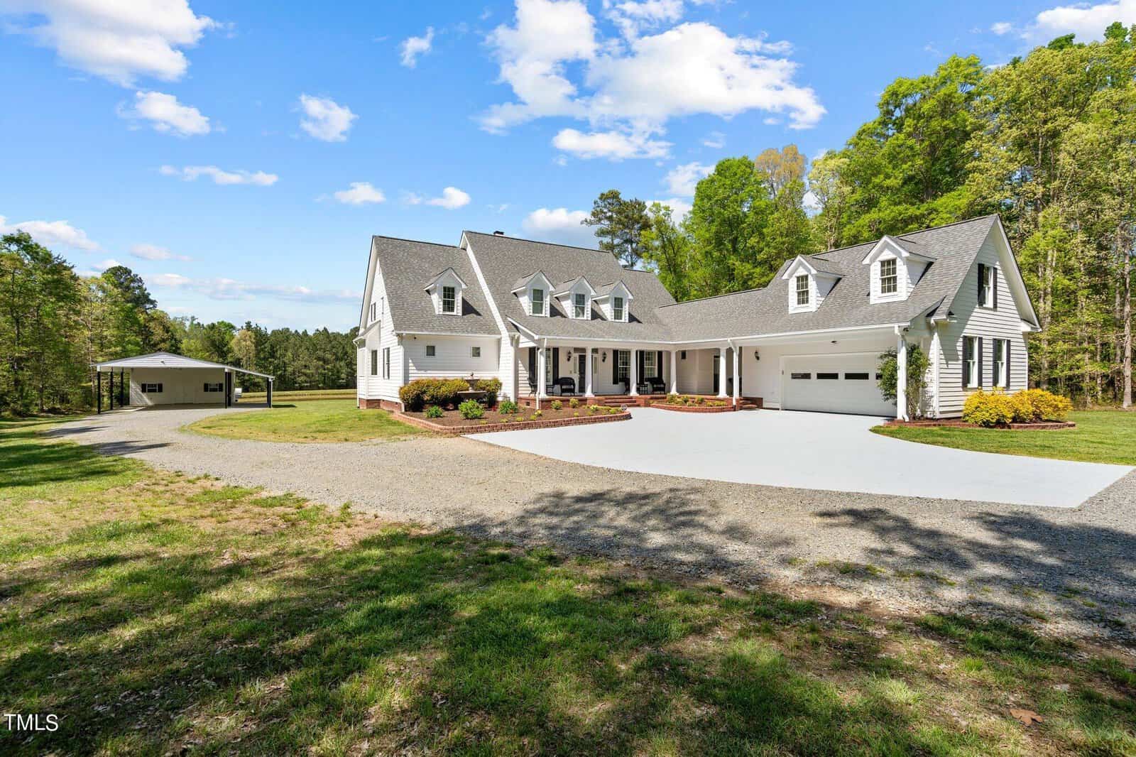 A large white house with a gray roof sits amidst a lush green landscape. It features multiple gables and a spacious driveway. A separate smaller structure is visible in the background, surrounded by trees under a blue sky with scattered clouds.