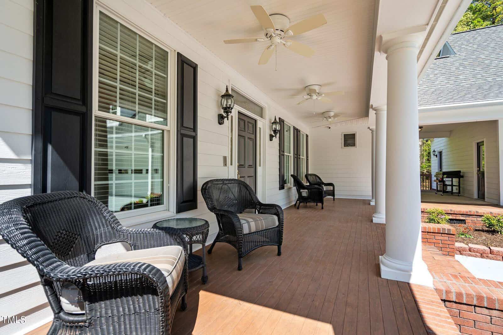 A spacious front porch with white siding and wooden flooring. It features black wicker chairs with cushions, ceiling fans, and decorative columns. The area is well-lit with outdoor wall lanterns and bordered by green foliage.