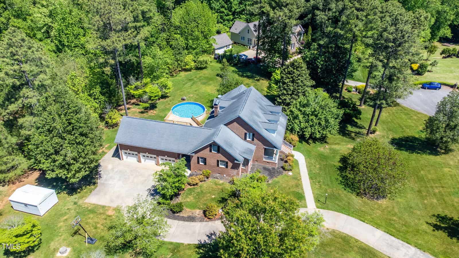 Aerial view of a large brick house surrounded by tall trees and greenery. The property features a circular above-ground pool in the backyard and a long driveway leading to a spacious garage. Neighboring homes are visible in the lush landscape.
