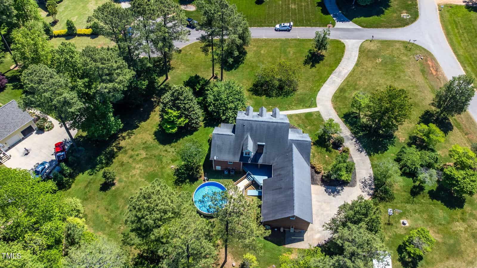 Aerial view of a large house with a gray roof surrounded by trees and green lawns. A round blue swimming pool is situated in the backyard. A curved driveway leads to the house, with several cars parked nearby. Other houses are visible in the vicinity.