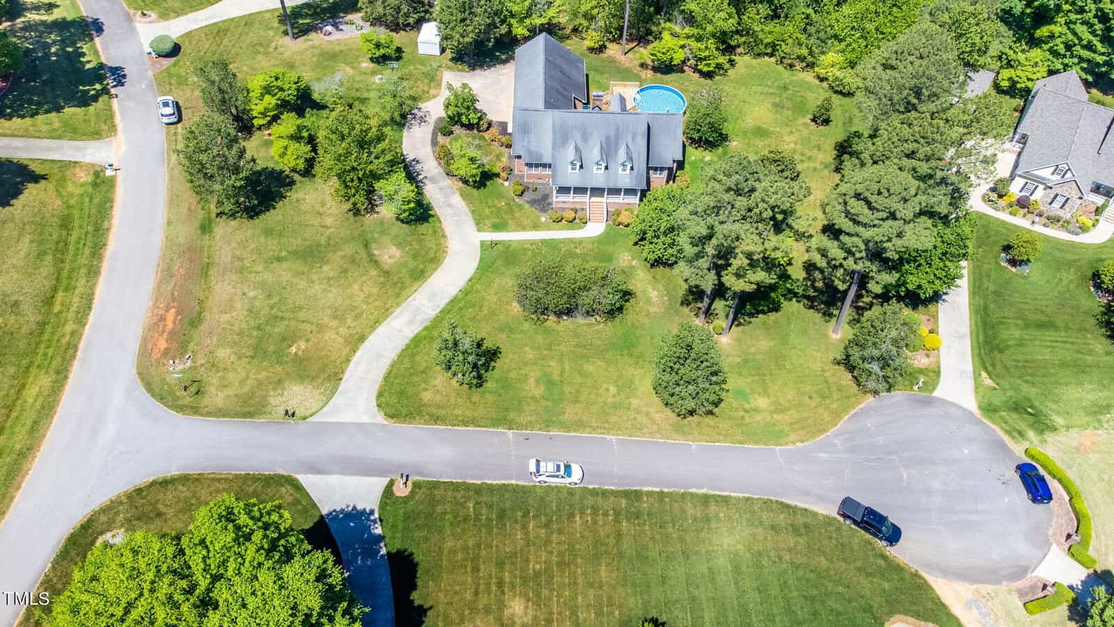 Aerial view of a suburban neighborhood featuring a large house with a swimming pool. The house is surrounded by trees and a well-maintained lawn. A few cars are parked on the street, and a curved driveway leads to the house.