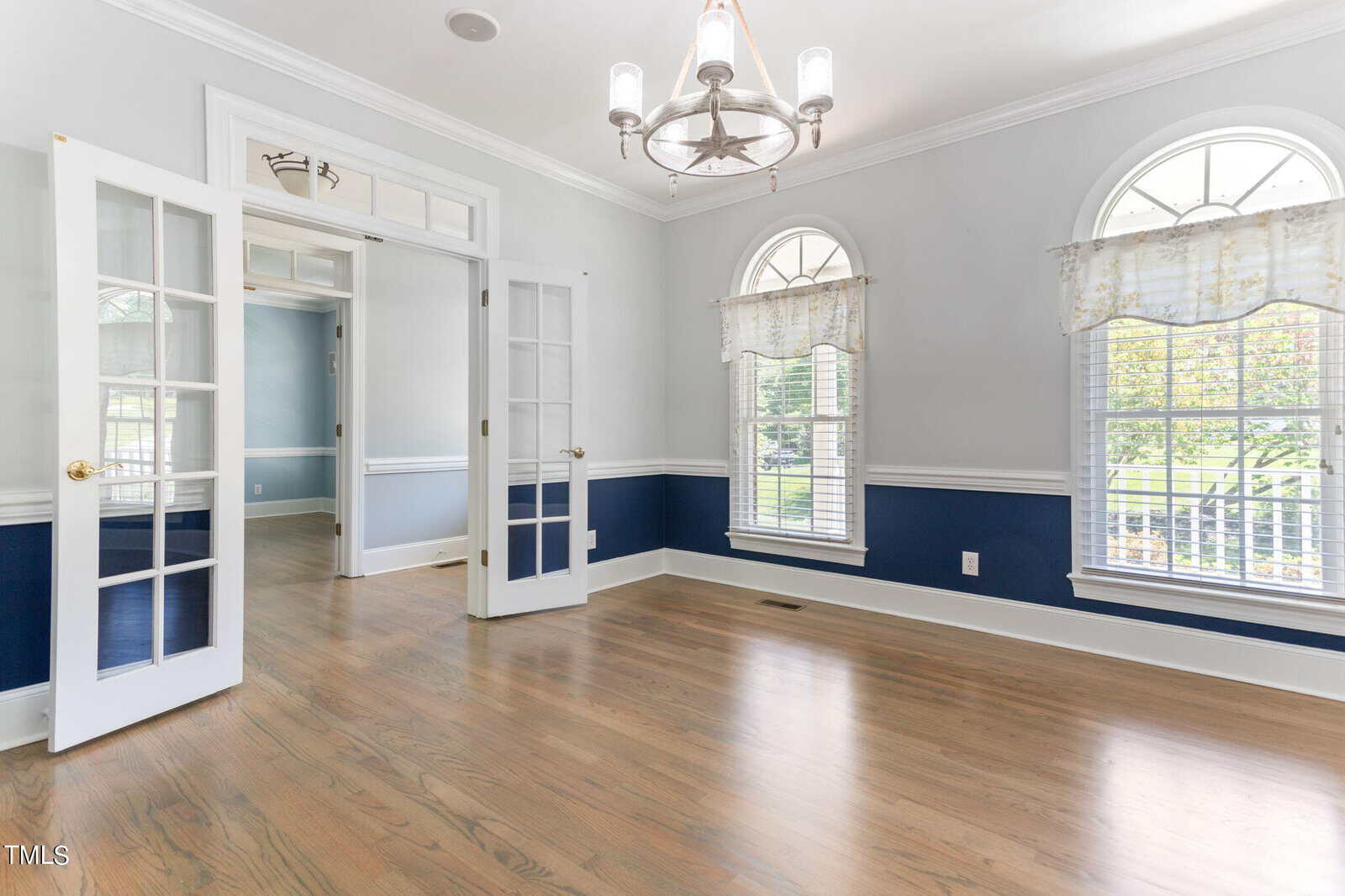 A bright, empty room featuring hardwood floors, white and blue walls, and arched windows with floral curtains. A chandelier hangs from the ceiling. French doors open to another room with similar decor.