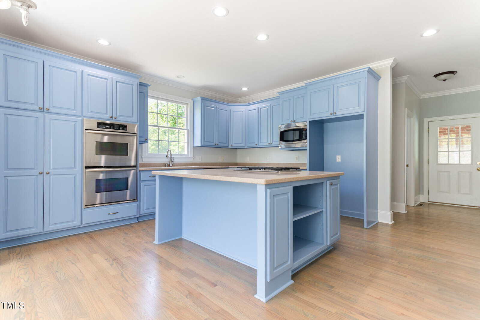 A spacious kitchen featuring light blue cabinetry, a central island with a stovetop, stainless steel double ovens, and a microwave. The room is brightened by natural light from a window above the sink. Hardwood floors and recessed lighting complete the look.