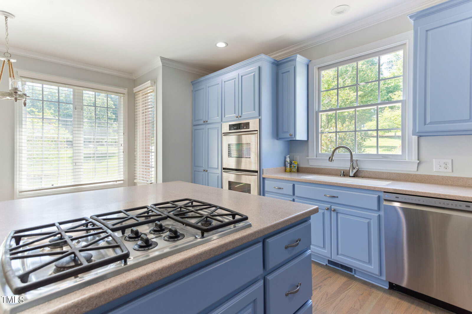 A bright kitchen with light blue cabinets, a double oven, and a gas stove on a central island. Large windows provide natural light, and a dishwasher is next to the sink under the window. Light wood flooring completes the airy feel.