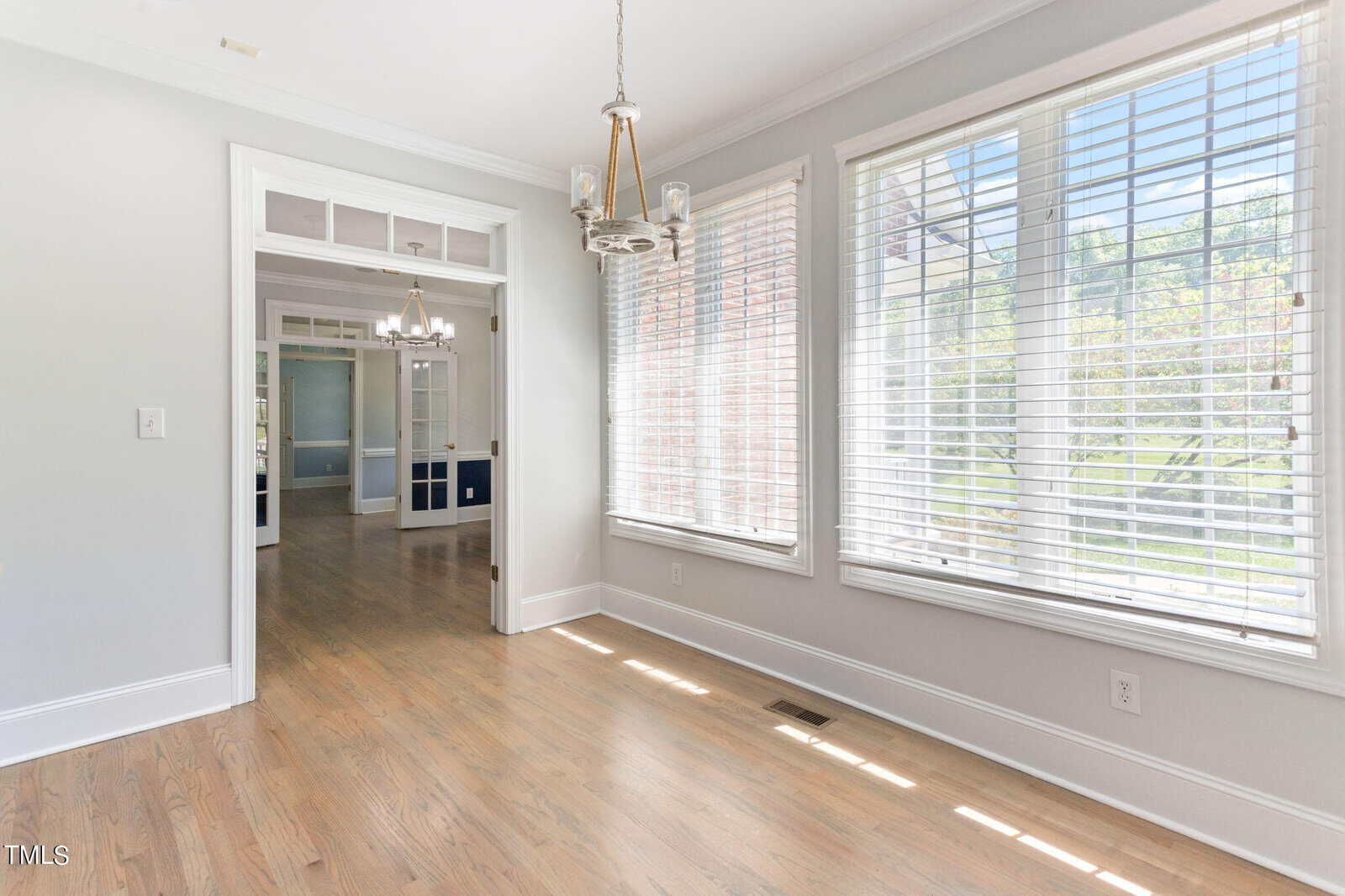 A bright, empty room with light hardwood floors and large windows with blinds, allowing natural light to flood in. The room is decorated with modern light fixtures and opens into another room with double doors.
