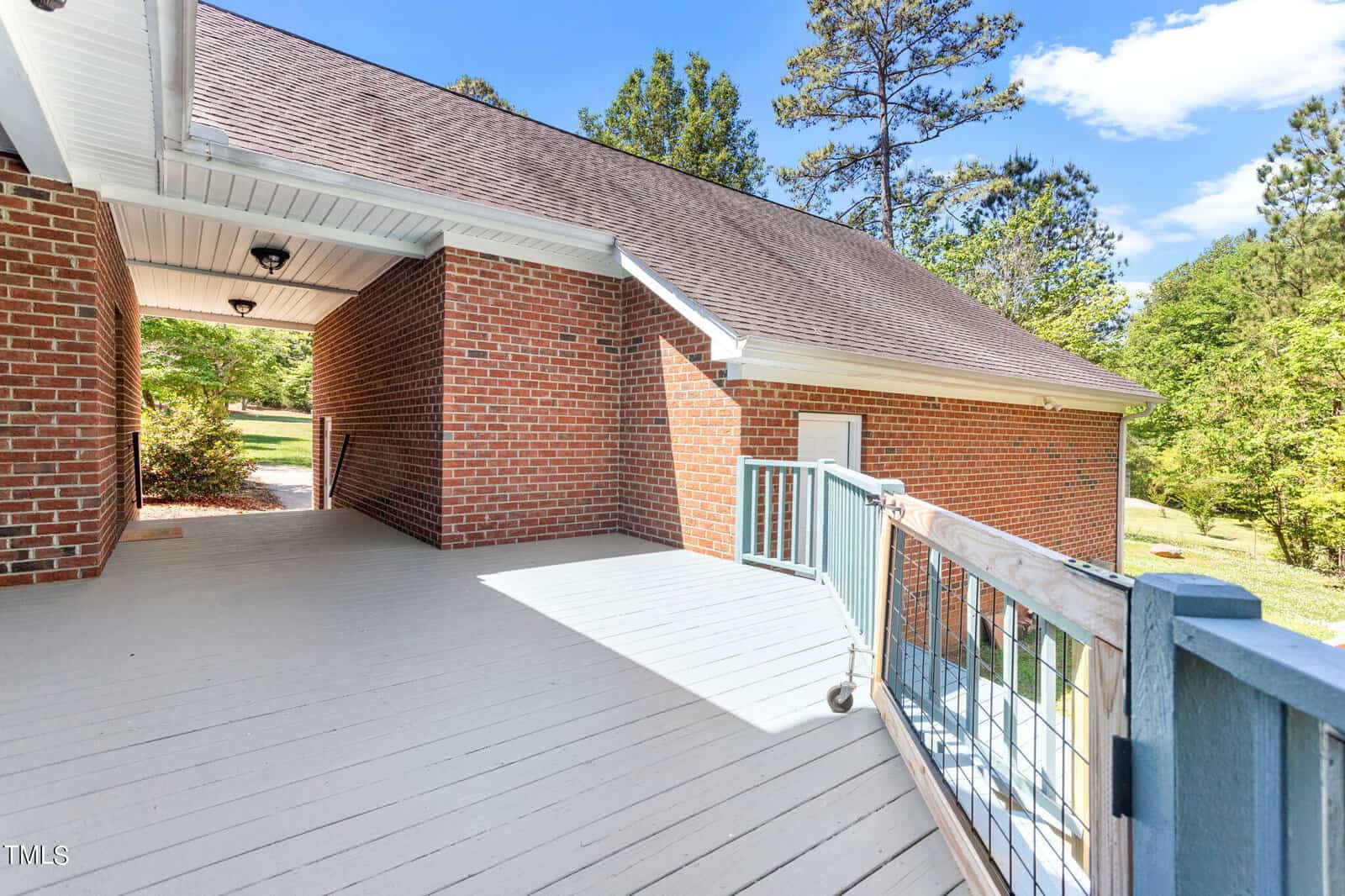 A spacious wooden deck attached to a red brick house with a sloped roof. The deck features a wooden railing and overlooks a lush, green backyard with tall trees under a bright blue sky.