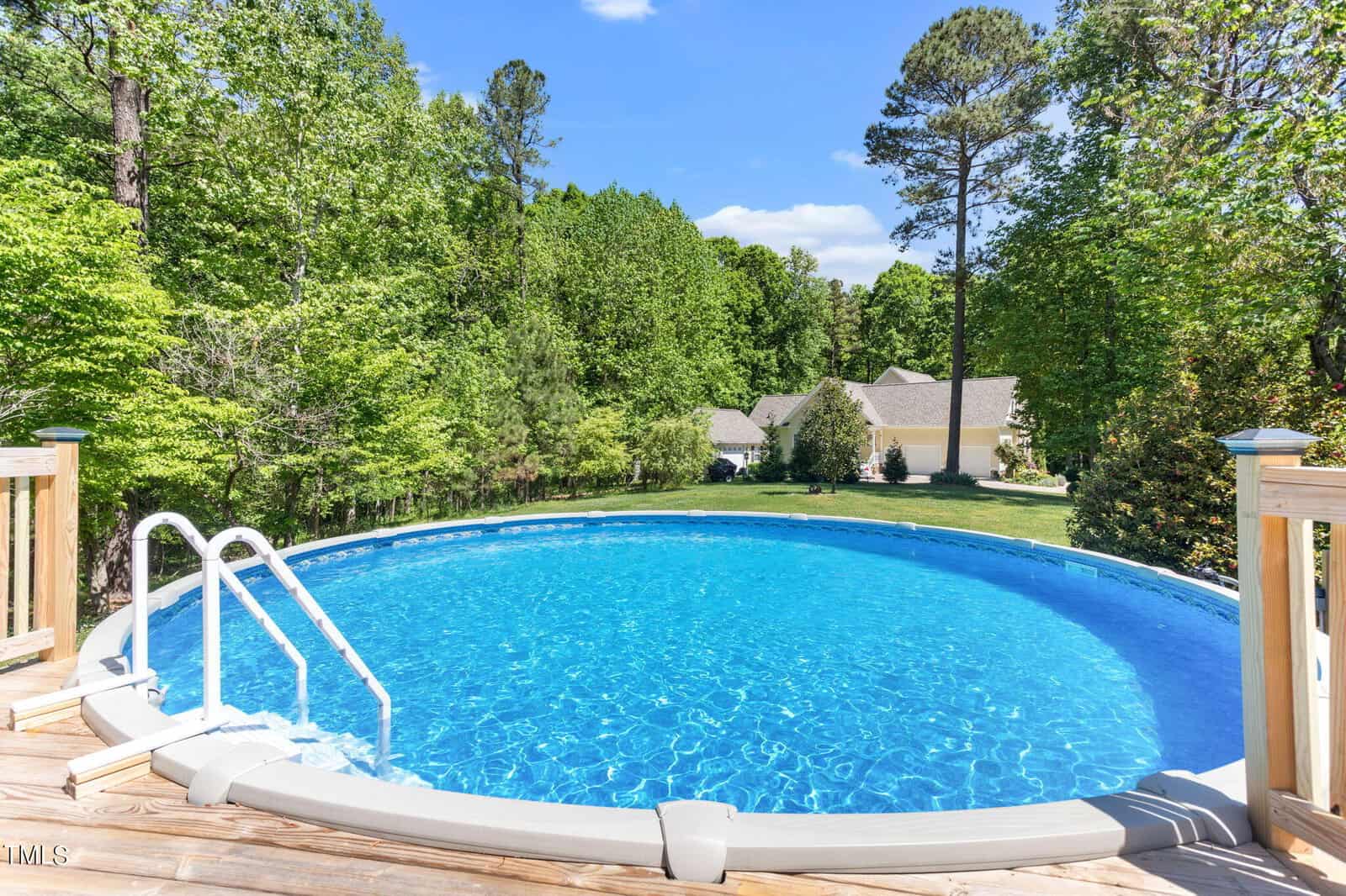 A round above-ground pool with clear blue water, surrounded by a wooden deck with metal stairs. Tall green trees and a white house with a sloped roof are visible in the background under a blue sky.