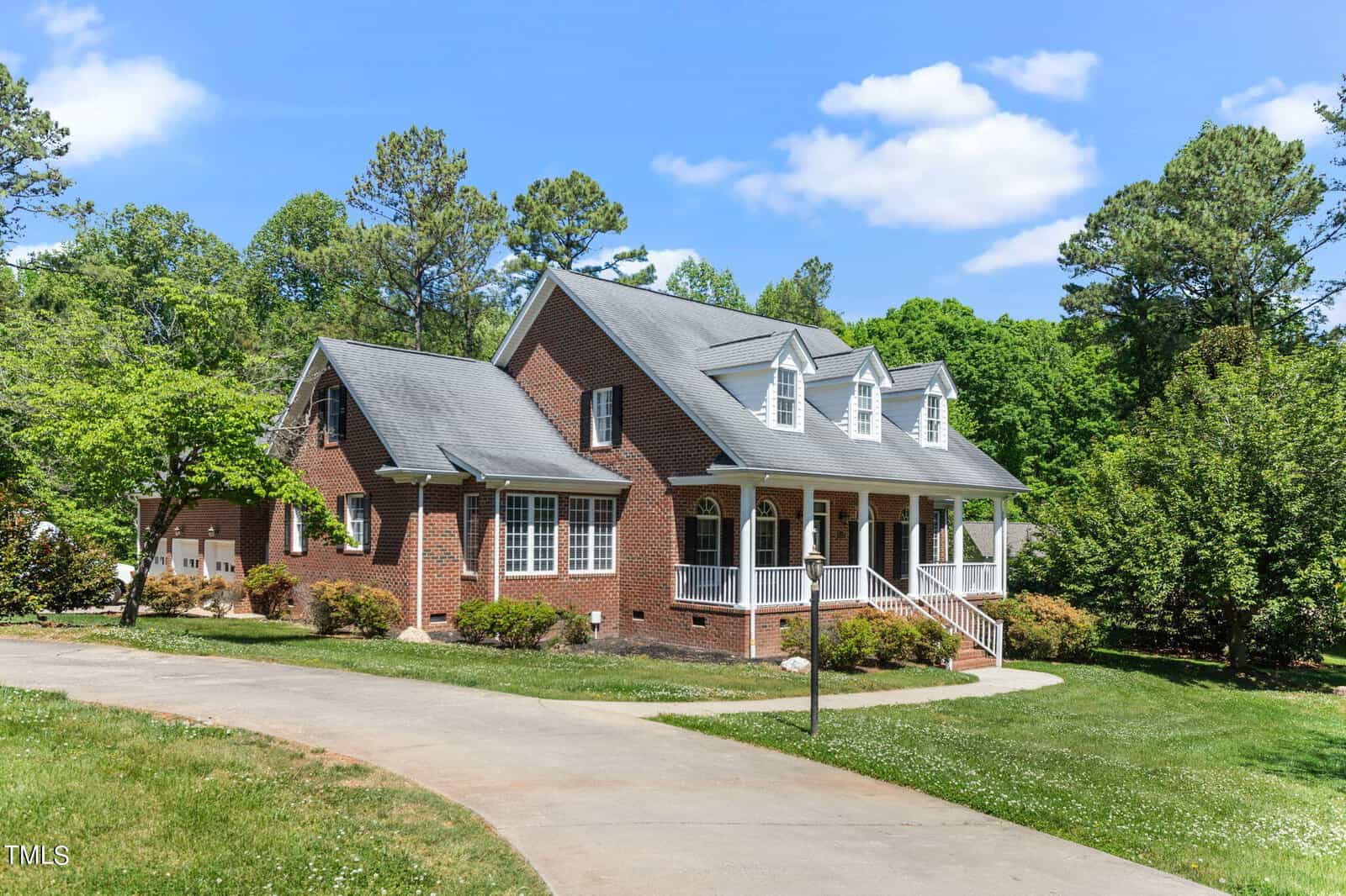 Nestled at 4139 Mistletoe Lane, this charming brick house with white trim boasts a welcoming covered porch, dormer windows, and a large front yard. Surrounded by lush green trees and set along a curved driveway, it lies under Oxford's bright blue sky dotted with fluffy clouds.