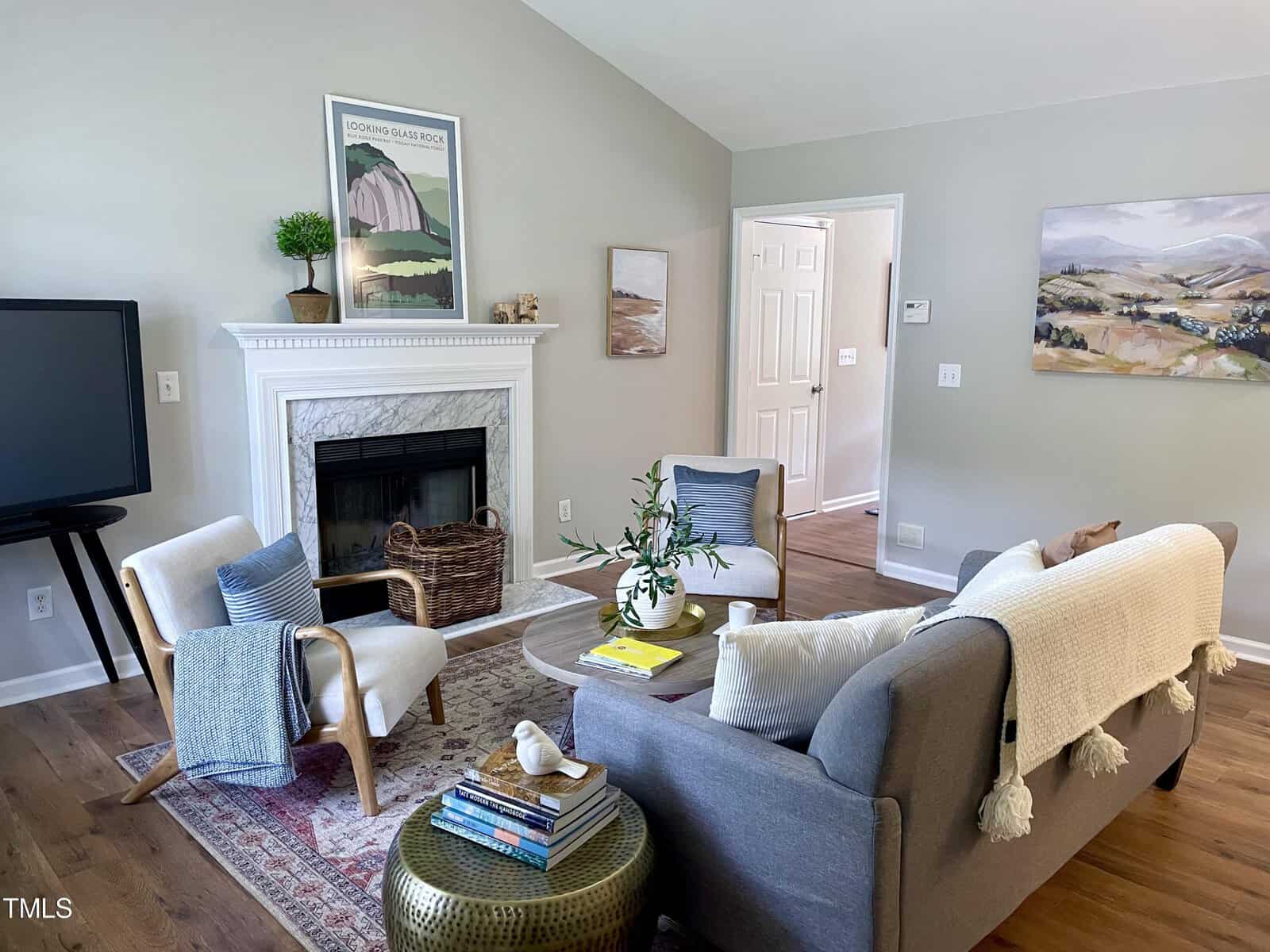 A cozy living room with a gray couch, two armchairs, and a TV on a stand. A fireplace with a marble surround is decorated with plants and artwork. A woven basket and a small table with books and a plant sit nearby. Light wood flooring completes the space.