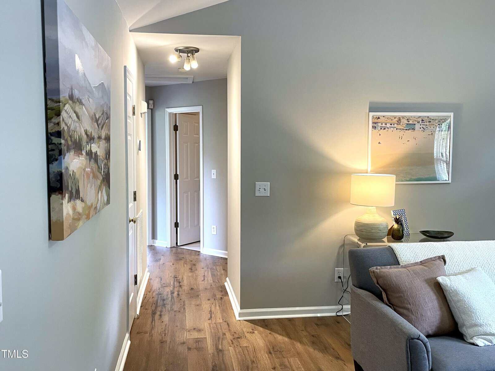 A cozy living room with gray walls and a hardwood floor. A framed painting hangs on one wall above a gray couch with cushions. A lamp on a side table casts a warm light, and an open doorway leads to a hallway.