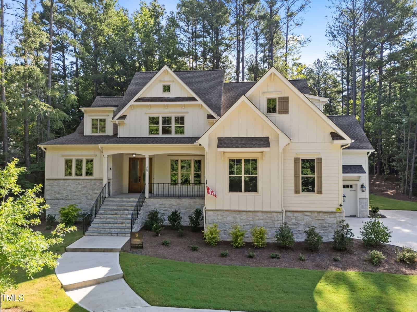 A large, two-story house with cream-colored siding and stone accents is surrounded by trees. The front entrance has steps, a covered porch, and a small American flag. A driveway curves around landscaped bushes and lawn.