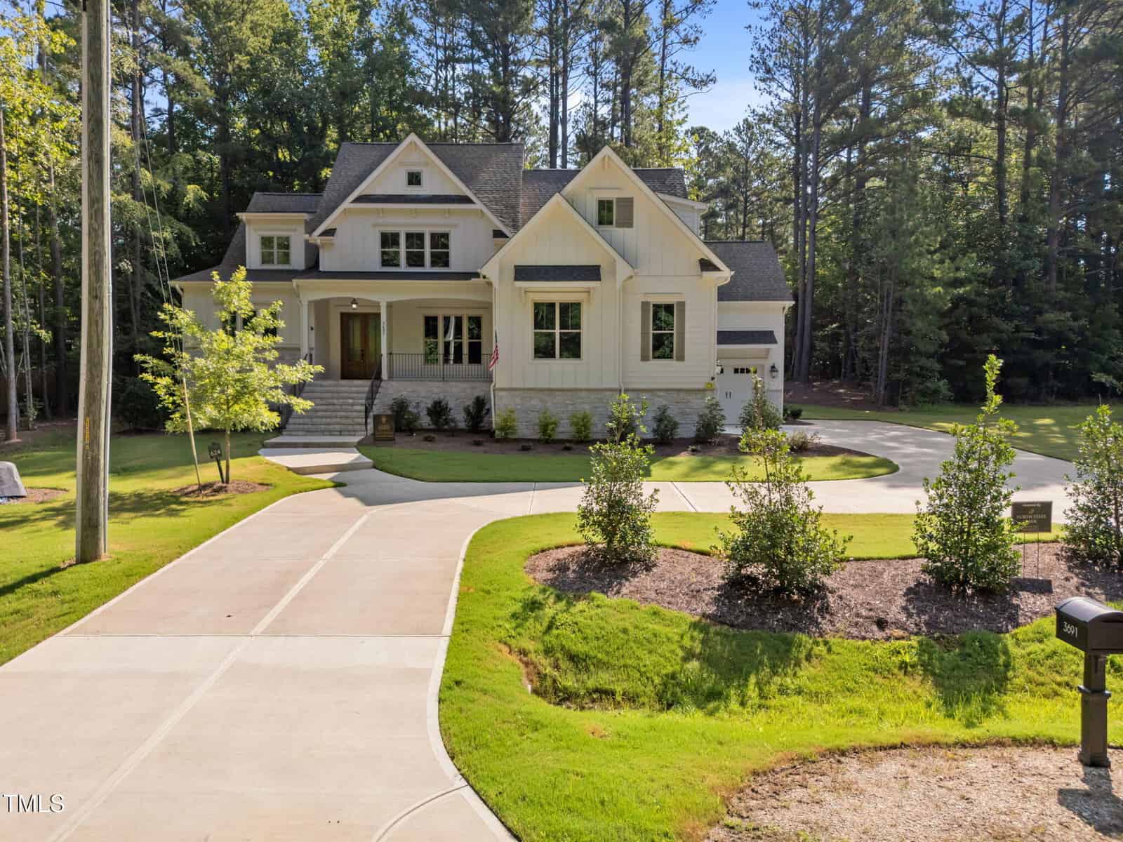 A large two-story house with white siding, multiple gables, and a covered front porch. The home has a circular driveway, manicured lawn, shrubs, and is surrounded by tall trees. A mailbox is visible in the foreground.
