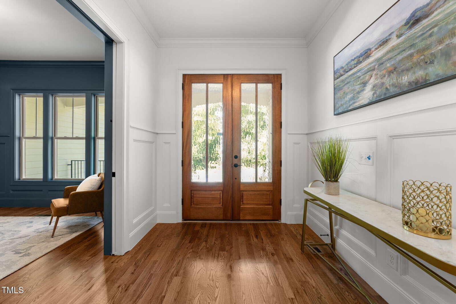 A bright entryway with double wooden front doors, white paneled walls, and a wooden floor. A narrow console table with decor sits to the right, and an adjacent room with blue walls and a brown chair is visible on the left.