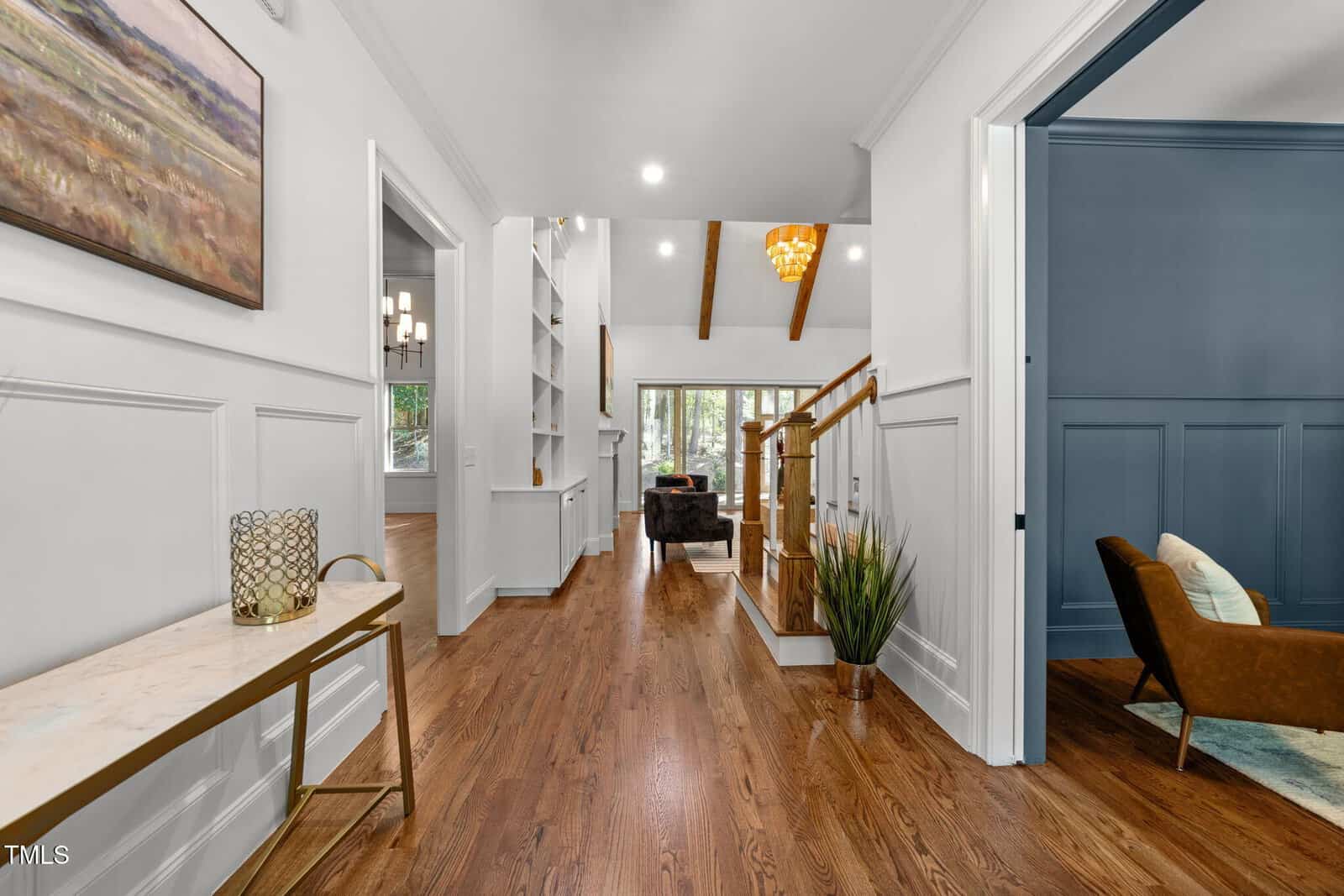 A bright hallway with hardwood floors, white walls, and wood ceiling beams leads to a living room. A blue accent room and a staircase are on the right; a console table and wall art are on the left.
