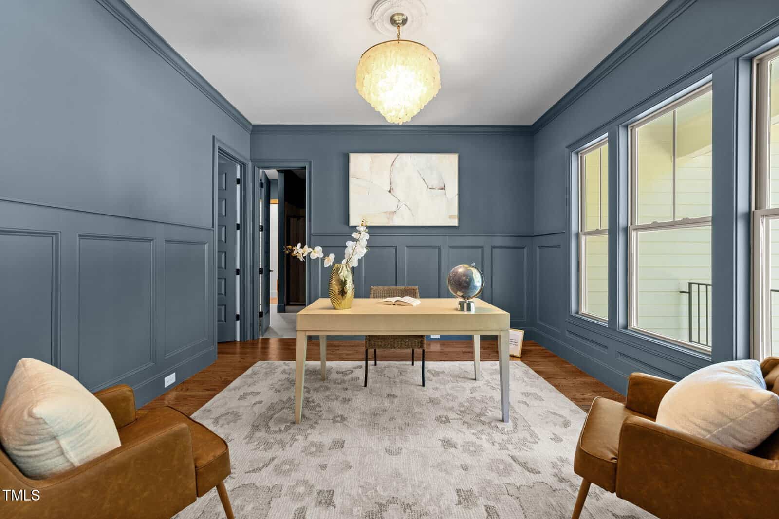 A home office with blue paneled walls, a light wood desk, a globe, a book, and a white orchid in a vase. Two brown armchairs with light pillows and a modern chandelier are also visible. Large windows line one wall.