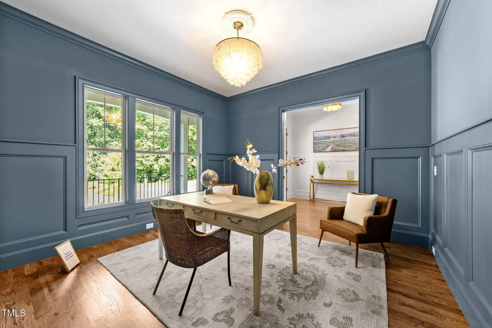 A home office with blue paneled walls, a beige desk, two chairs, a light-colored rug, and a chandelier. A window lets in natural light, and there are decorative items on the desk and console table in the background.