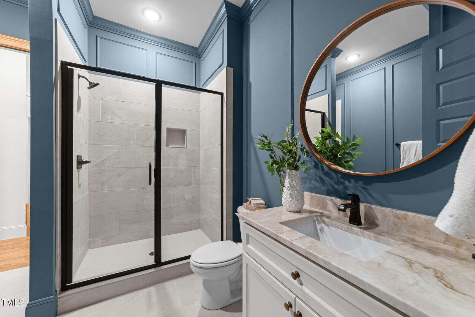 Modern bathroom with blue paneled walls, a glass-enclosed shower, white toilet, white vanity with marble countertop, round mirror, black fixtures, and a green plant in a white vase on the counter.