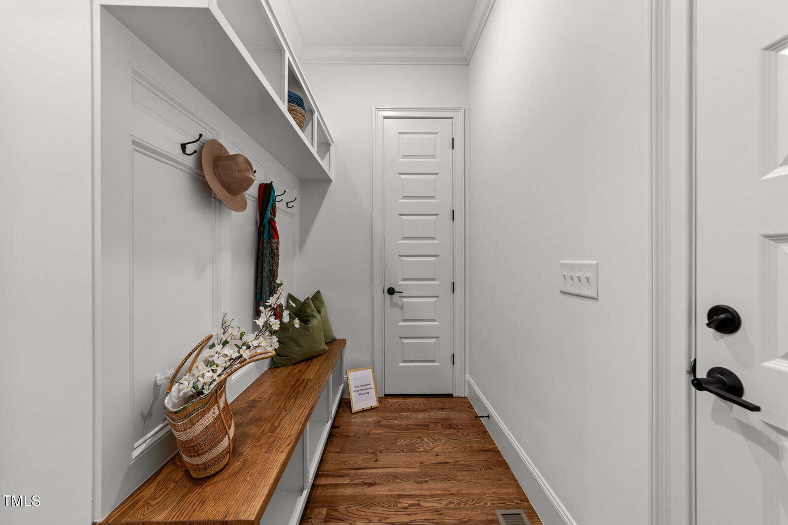 A hallway with white walls and a wooden bench, featuring hooks with hats and scarves, a green pillow, a wicker basket with flowers, and a framed quote. A white door with panels is at the end of the hallway.