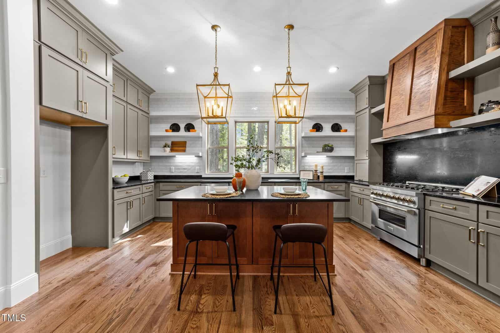 Modern kitchen with hardwood floors, gray cabinets, a wood-accented range hood, a central island with two chairs, two pendant lights, open shelves, and a window above the sink letting in natural light.