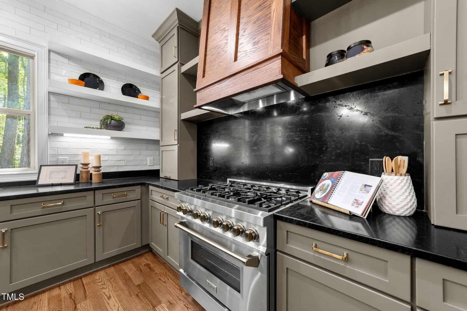 Modern kitchen with gray cabinets, black countertops, a stainless steel gas stove, wooden range hood, open shelves with decor, a cookbook stand, and hardwood floors. Sunlight streams in from a window on the left.