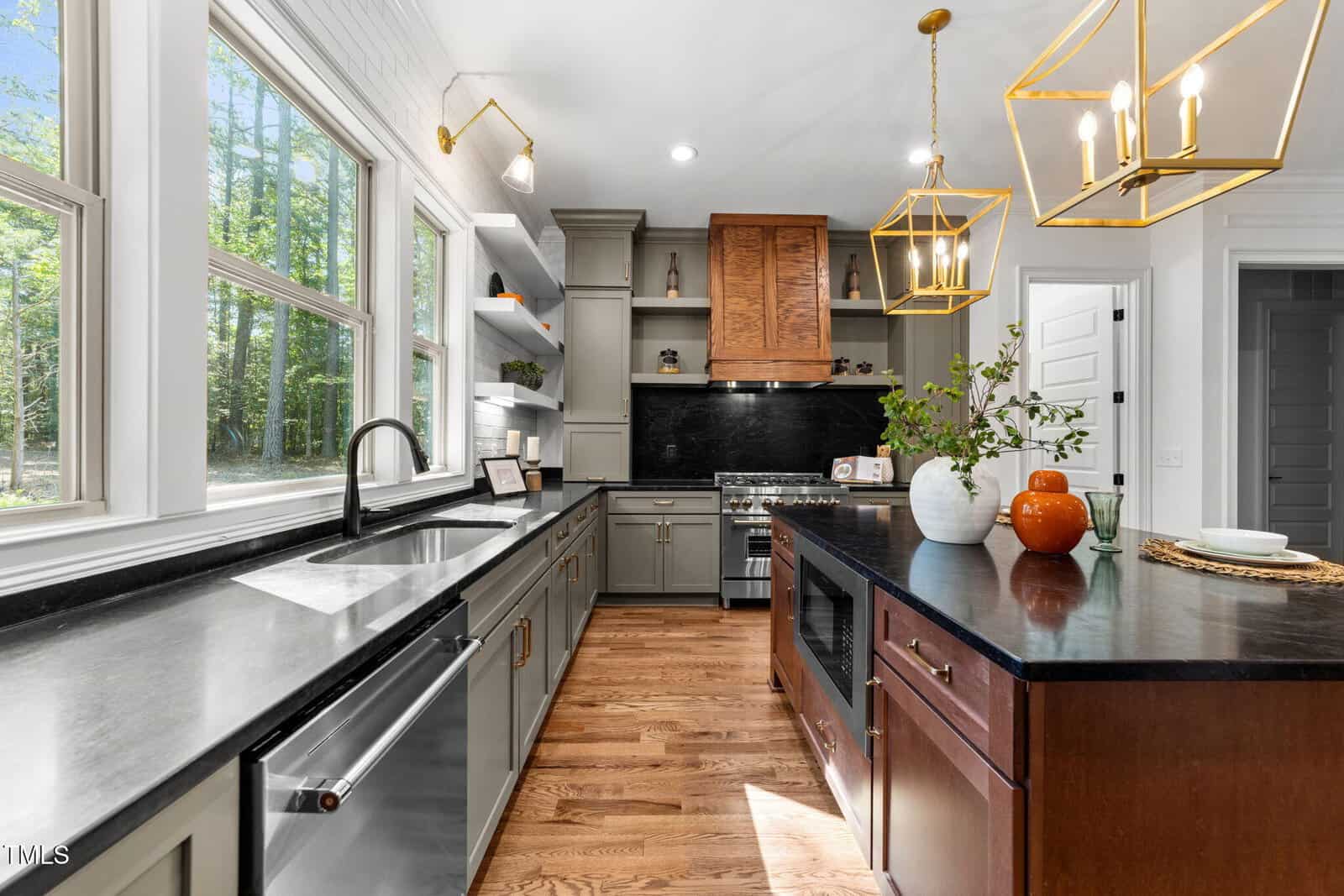 Modern kitchen with stainless steel appliances, large windows, gray cabinets, wood floors, black countertops, and gold pendant lights above a dark island with decor and a plant.