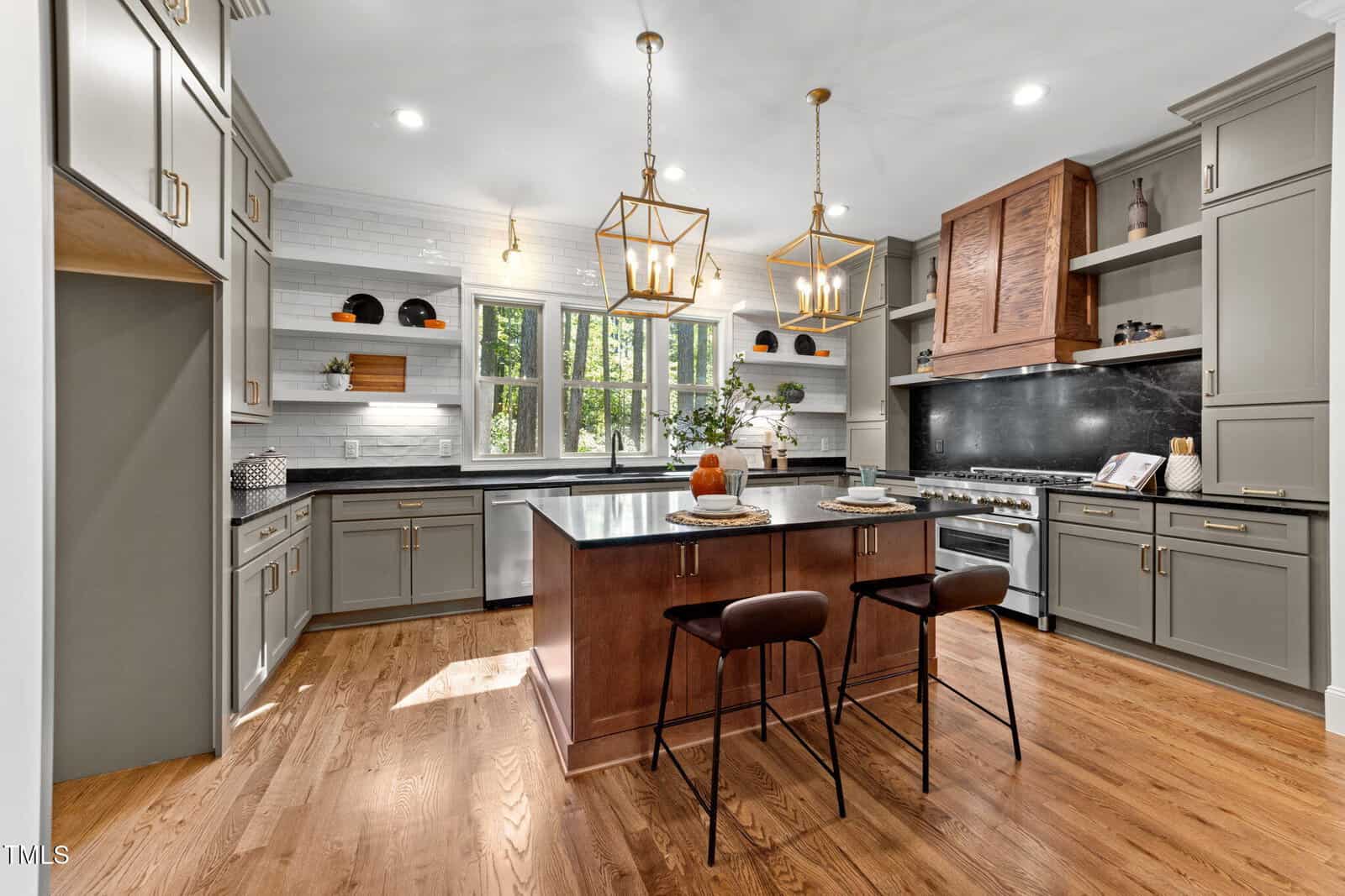 Modern kitchen with gray cabinets, a wooden island with two barstools, pendant lights, stainless steel appliances, open shelves, black backsplash, and large windows letting in natural light.