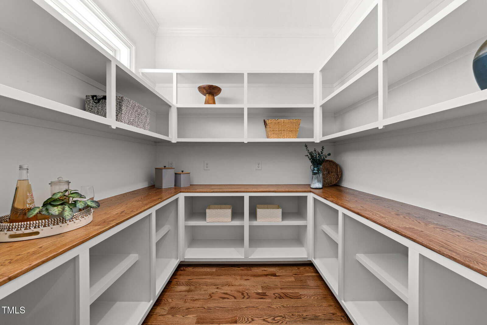 A spacious walk-in pantry with white shelves, wooden countertops, and decorative items including baskets, jars, a plant, and a tray with bottles on a hardwood floor.