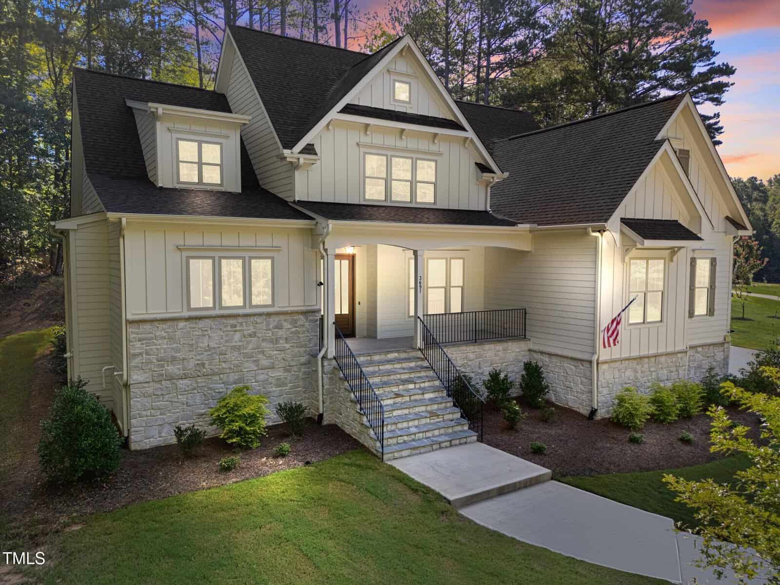 This two-story beige house with stone siding, black roof, and multiple gables sits at 3691 Graham Sherron Road in Wake Forest. A front porch leads to the entrance, while a landscaped lawn and sunset-lit trees complete the picturesque scene.