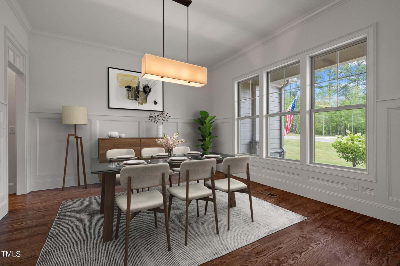 A modern dining room with a glass table set for six, beige chairs, and a rectangular pendant light. A large window shows trees and an American flag outside. The room features white walls and a wooden floor with a gray rug.