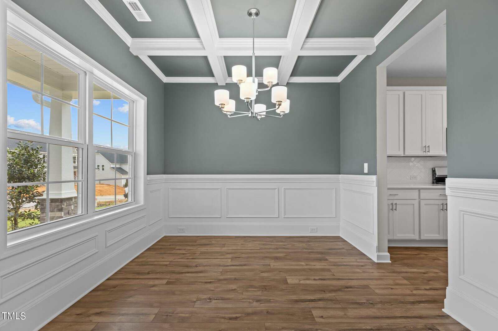 A bright dining room with gray walls, white wainscoting, coffered ceiling, wood flooring, large windows, a modern chandelier, and an open doorway to a white kitchen.